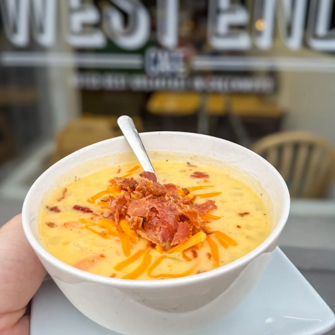A creamy bowl of soup topped with crispy bacon and shredded cheese, served against a backdrop of a cafe.