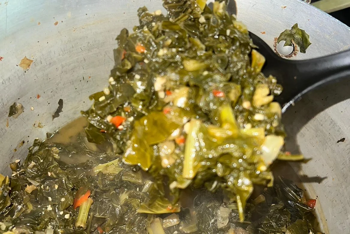 A close-up of cooked greens in a pot, featuring vibrant leafy vegetables with bits of seasoning visible. The dish appears flavorful and nutritious.