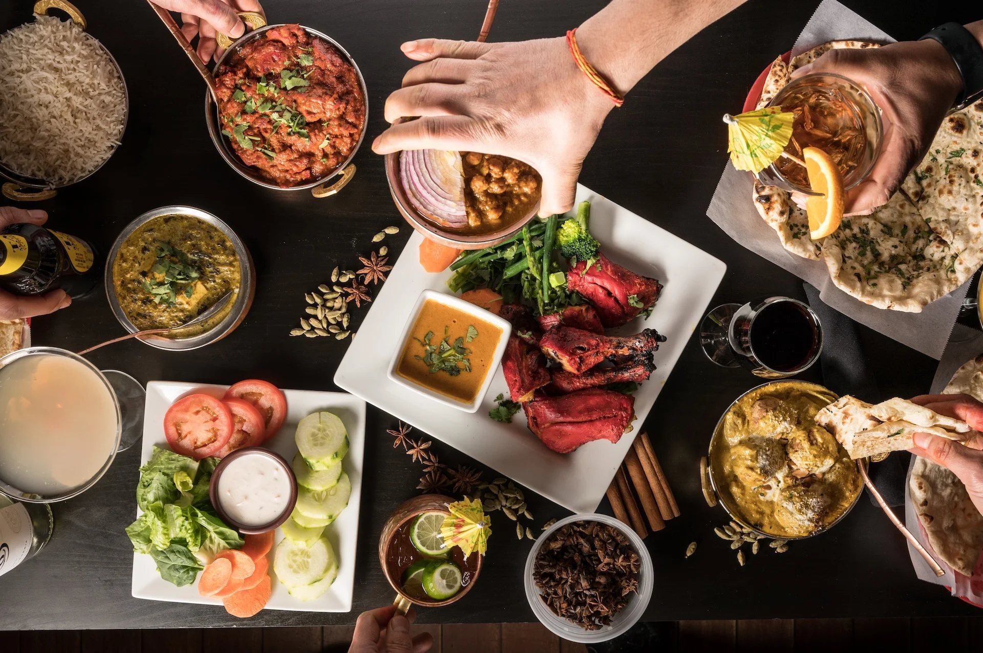 A vibrant, top-down view of an exceptional dining experience at Mint Indian Restaurant in Denver, with hands reaching to share a communal feast of tandoori chicken, curries, and naan bread.