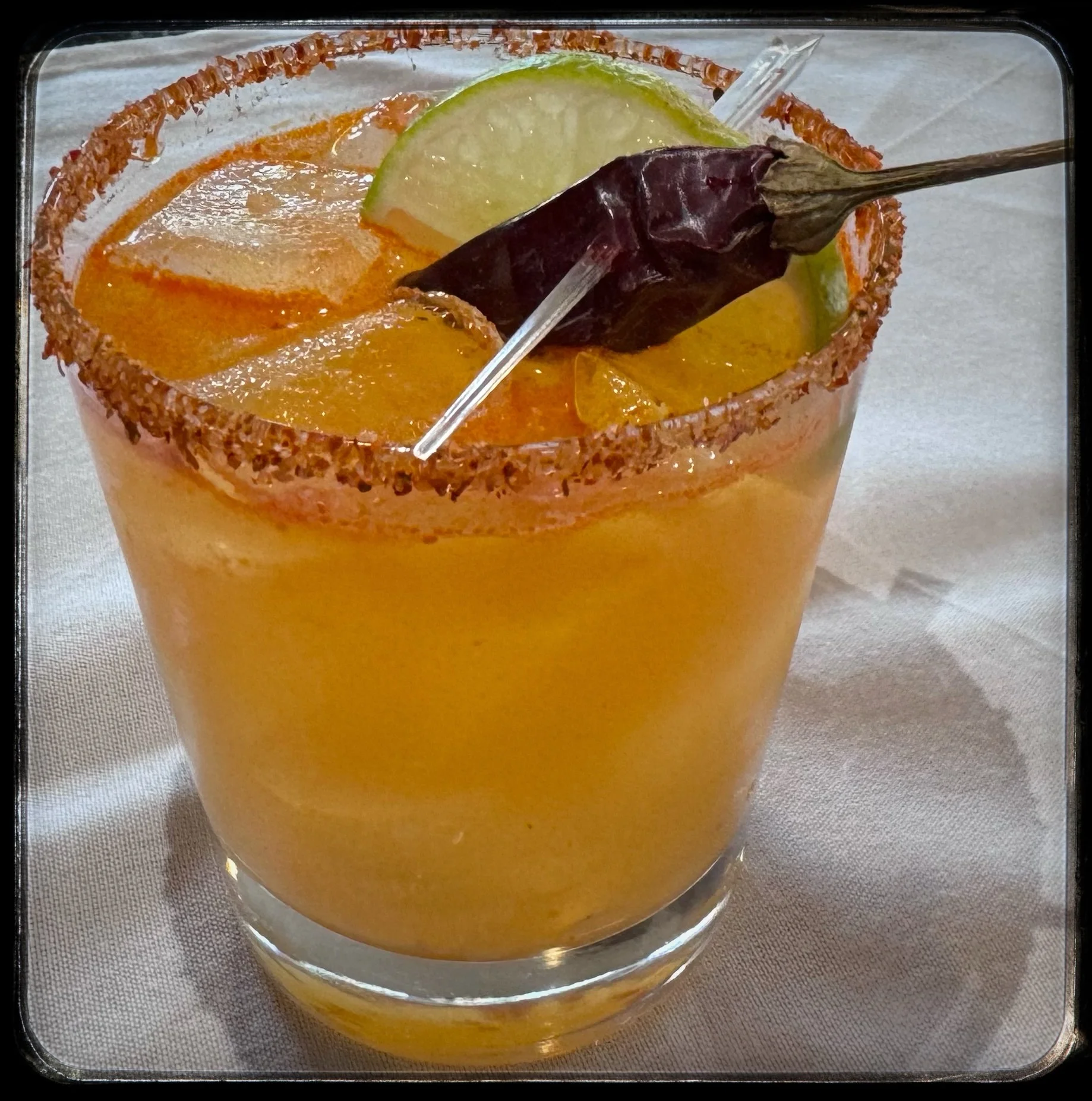 A vibrant orange cocktail in a glass with a chili powder-rimmed edge, garnished with lime slice and a dried chili pepper served at Toscana on Main