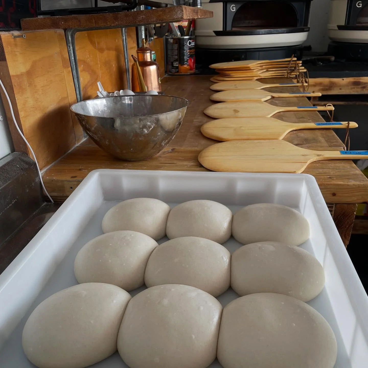 A tray of pizza dough balls resting, ready for use in cooking. In the background, wooden peels are lined up on a countertop.