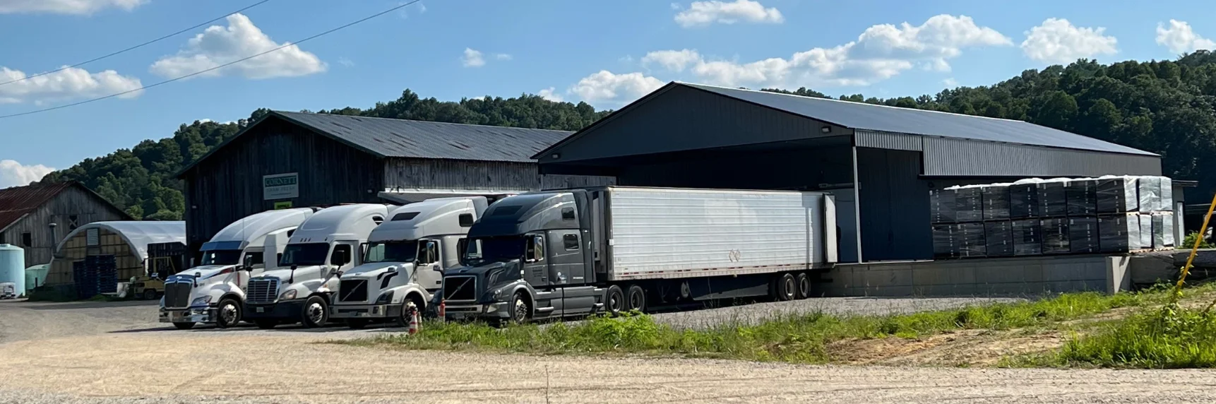Trucks parked at a rural warehouse under a blue sky with fluffy clouds