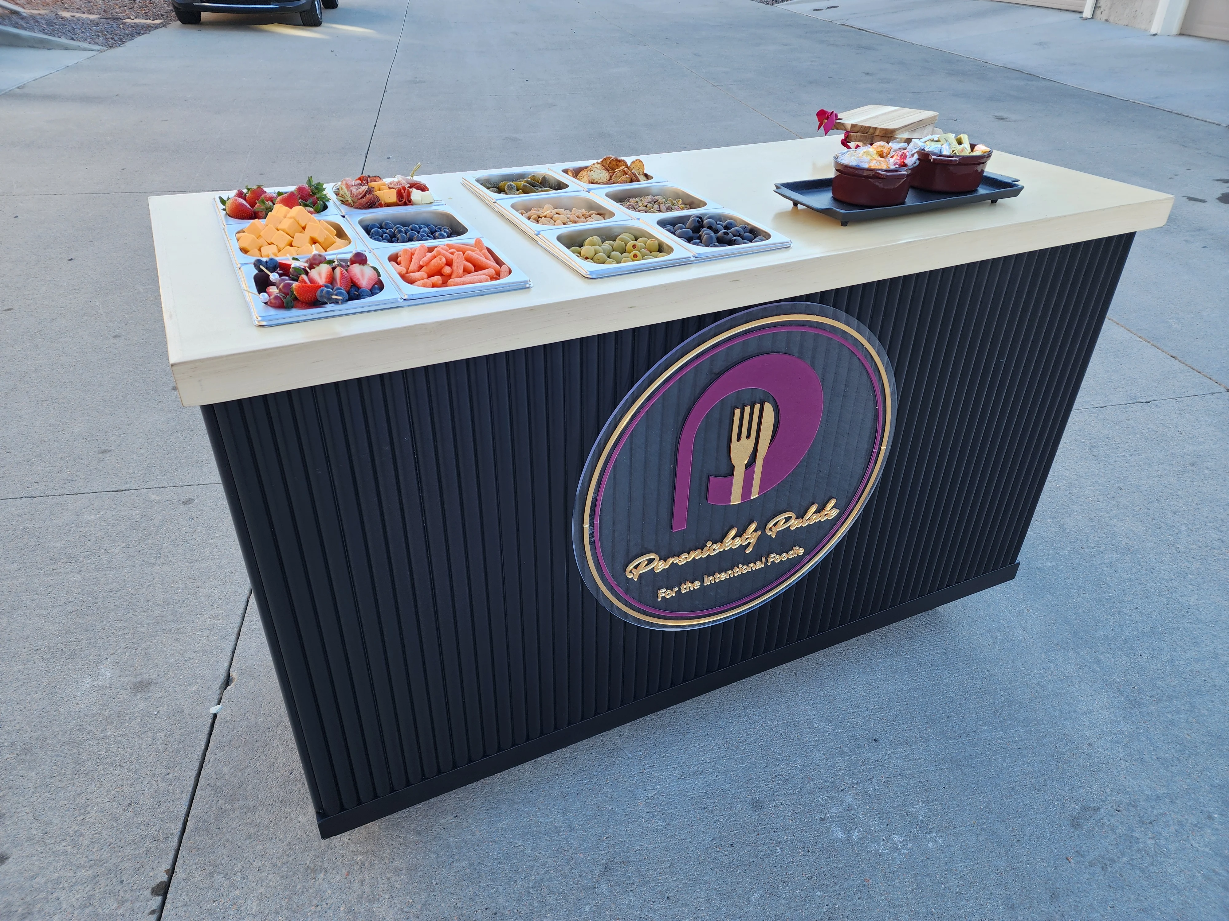 An outdoor food display featuring an assortment of colorful fruits, snacks, and cheeses on a modern serving table.