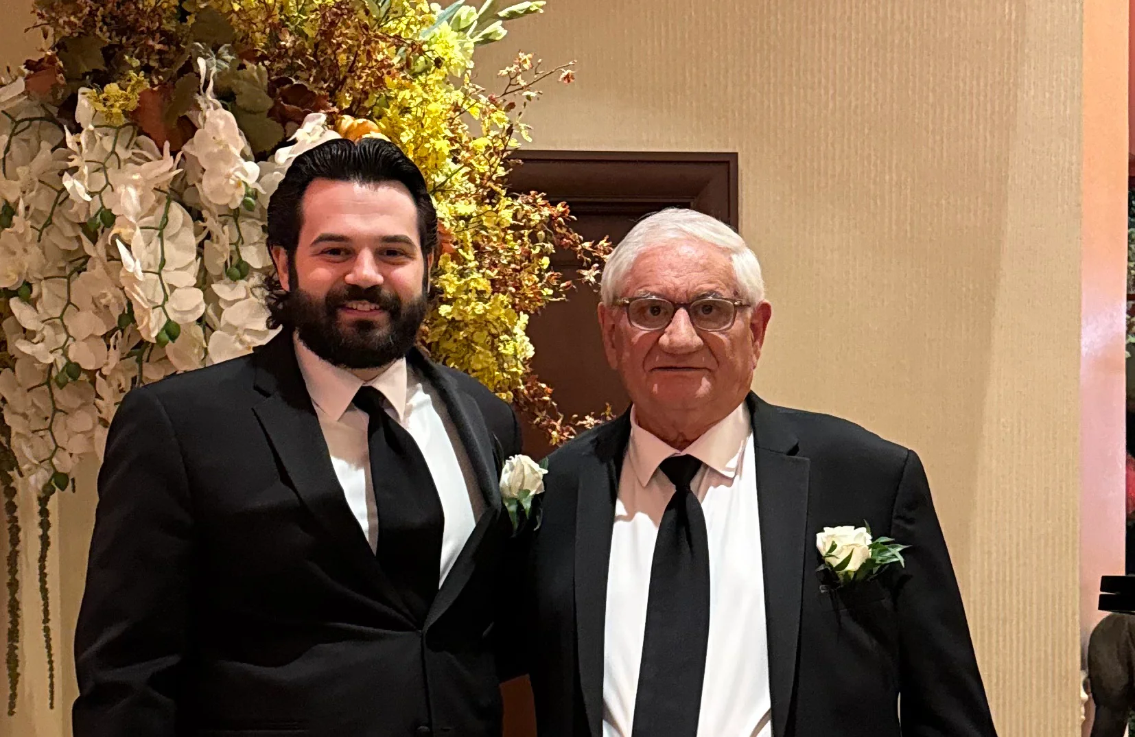 Formally dressed men posing inside an elegant restaurant beside floral arrangements and piano