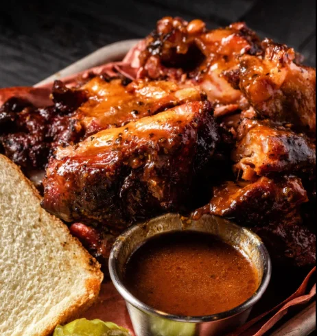 A plate of smoked meat garnished with barbecue sauce, accompanied by a slice of bread and a small bowl of additional sauce.