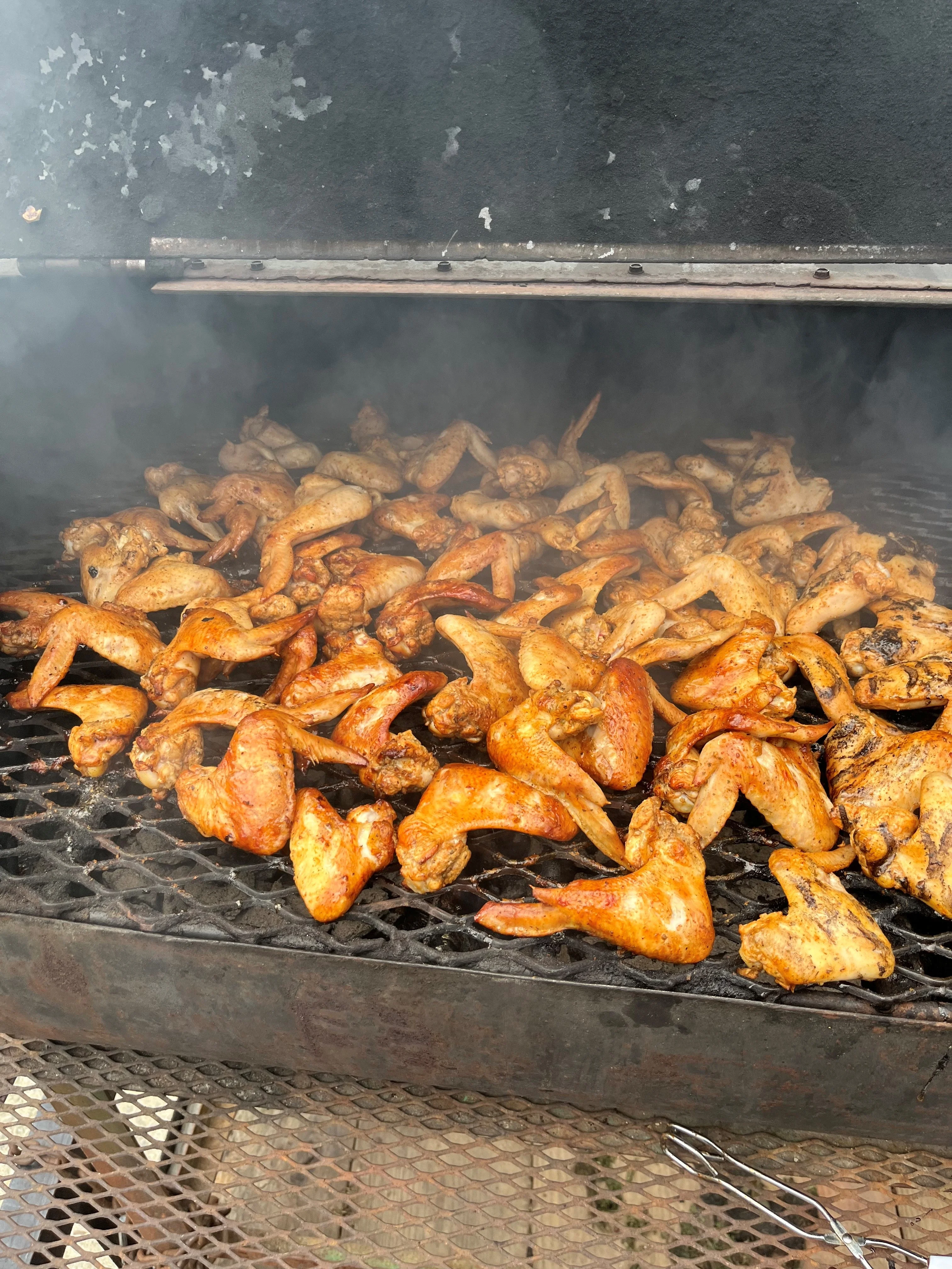 A large quantity of chicken wings being smoked on a grill. The wings are coated in spices and some are glistening from the cooking process.
