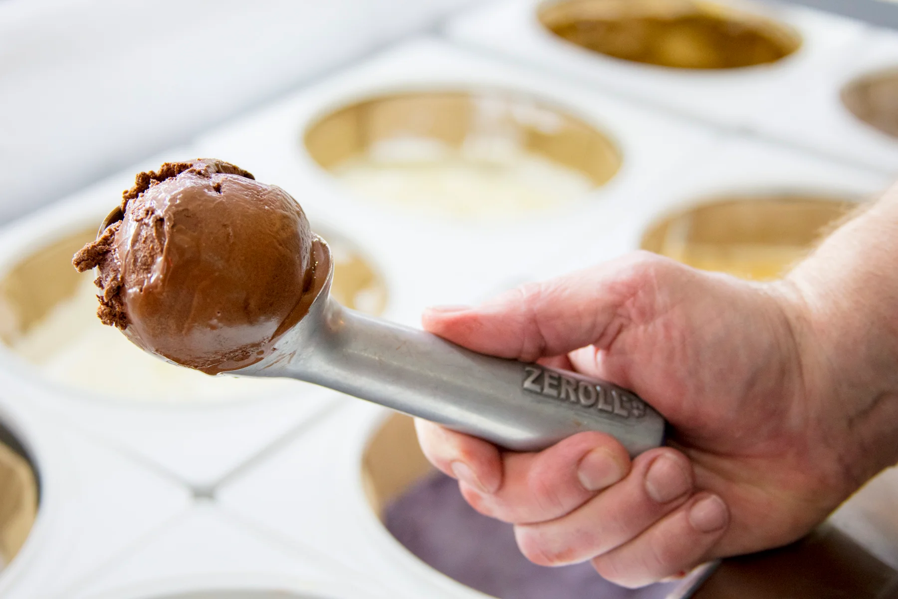 Hand holding an ice cream scooper with a rich, creamy chocolate scoop in front of a display of assorted ice cream tubs at a Northwest Arkansas ice cream shop.