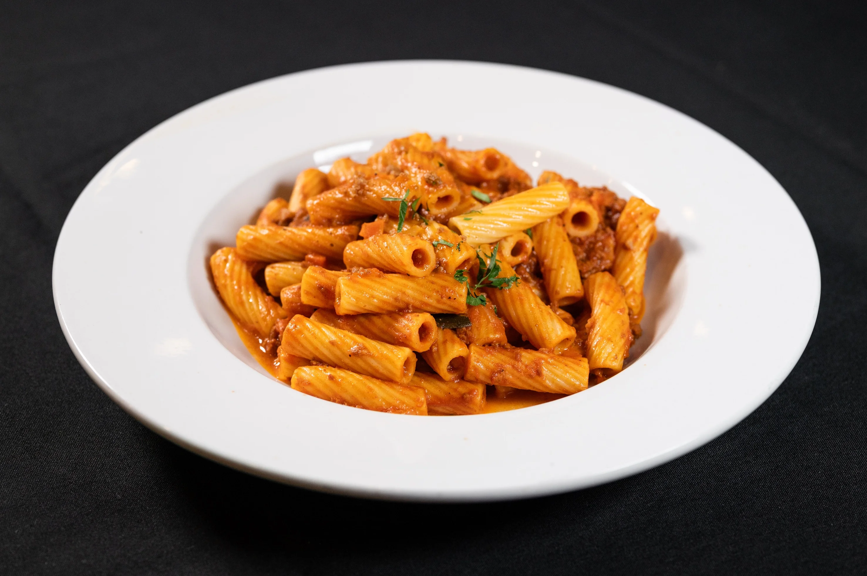 Plate of rigatoni pasta with rich red tomato sauce, garnished with fresh herbs, served on a white plate against a dark background. Warm and appetizing served at Toscana on Main