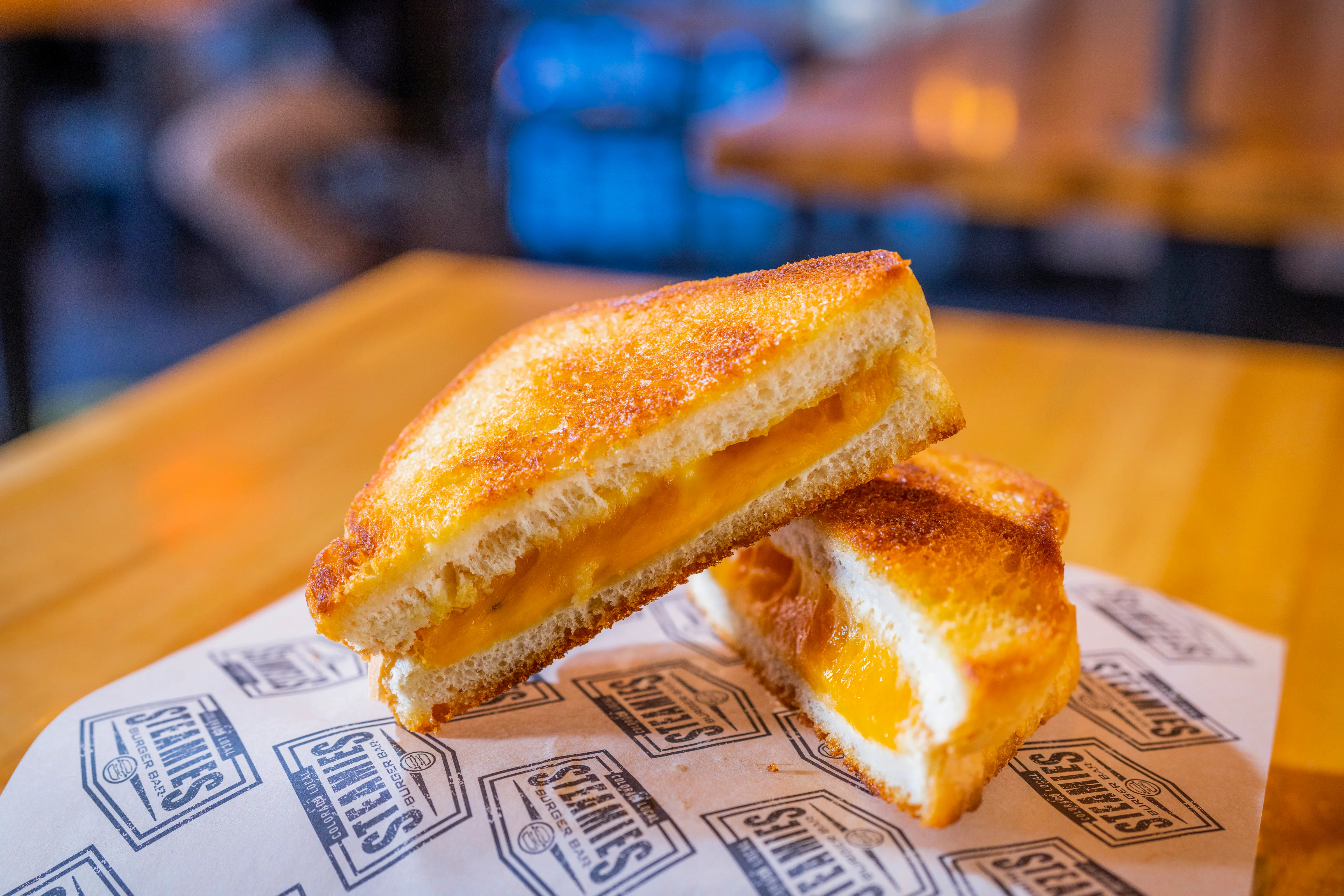 A grilled cheese sandwich cut in half, revealing melted cheese between two golden brown slices of bread, served on a branded paper.