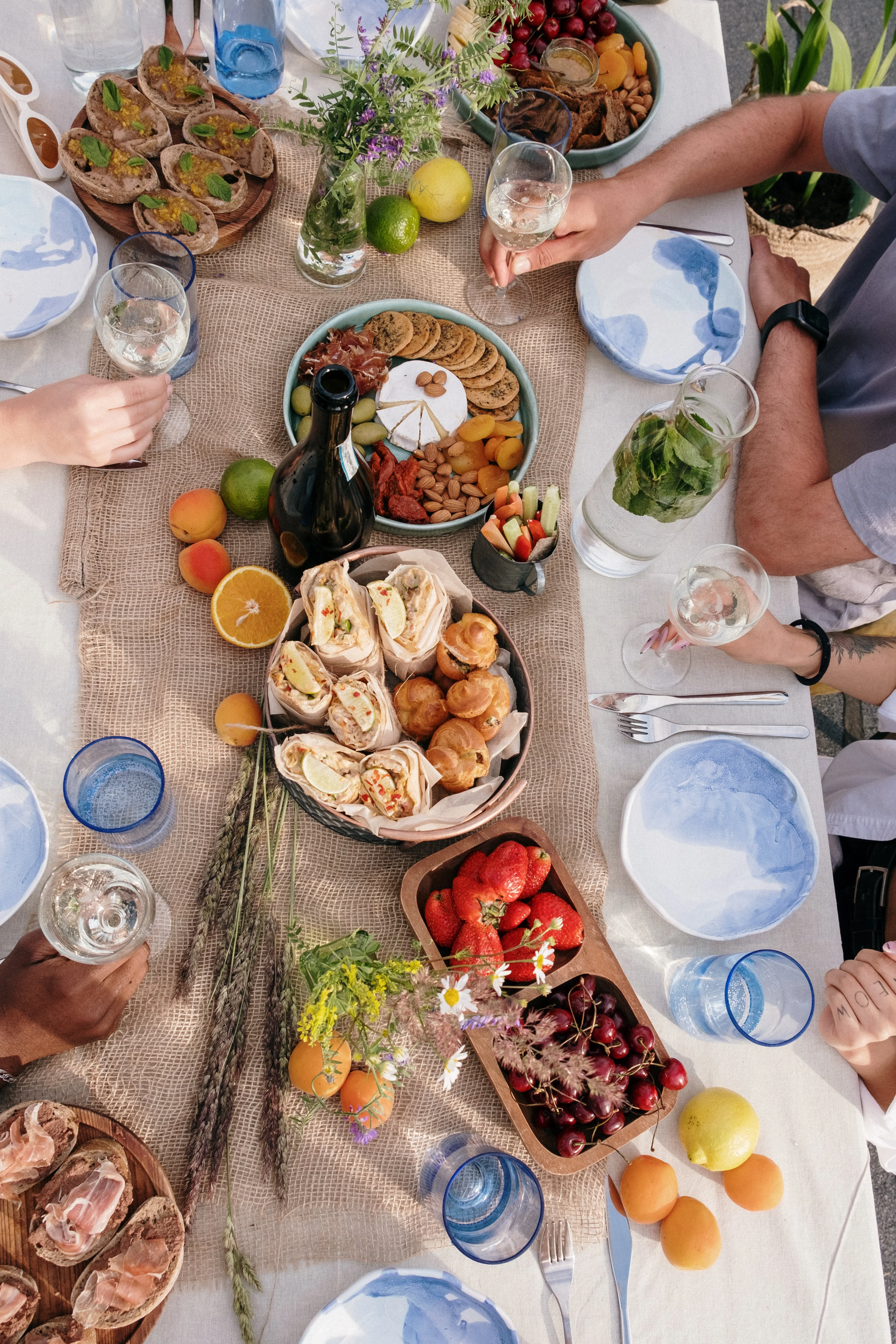 Overhead Mediterranean-style picnic spread with artisanal breads, pastries, fruit, cheese, and drinks
