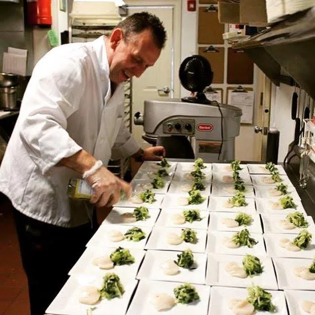 A chef in a professional kitchen meticulously plating food, adding finishing touches to several dishes arranged in a line. Ingredients are carefully presented.