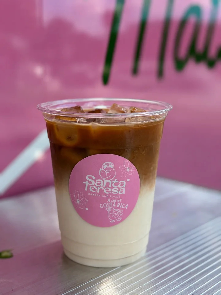 A layered iced beverage with coffee on top and cream at the bottom, served in a transparent cup with a pink logo.