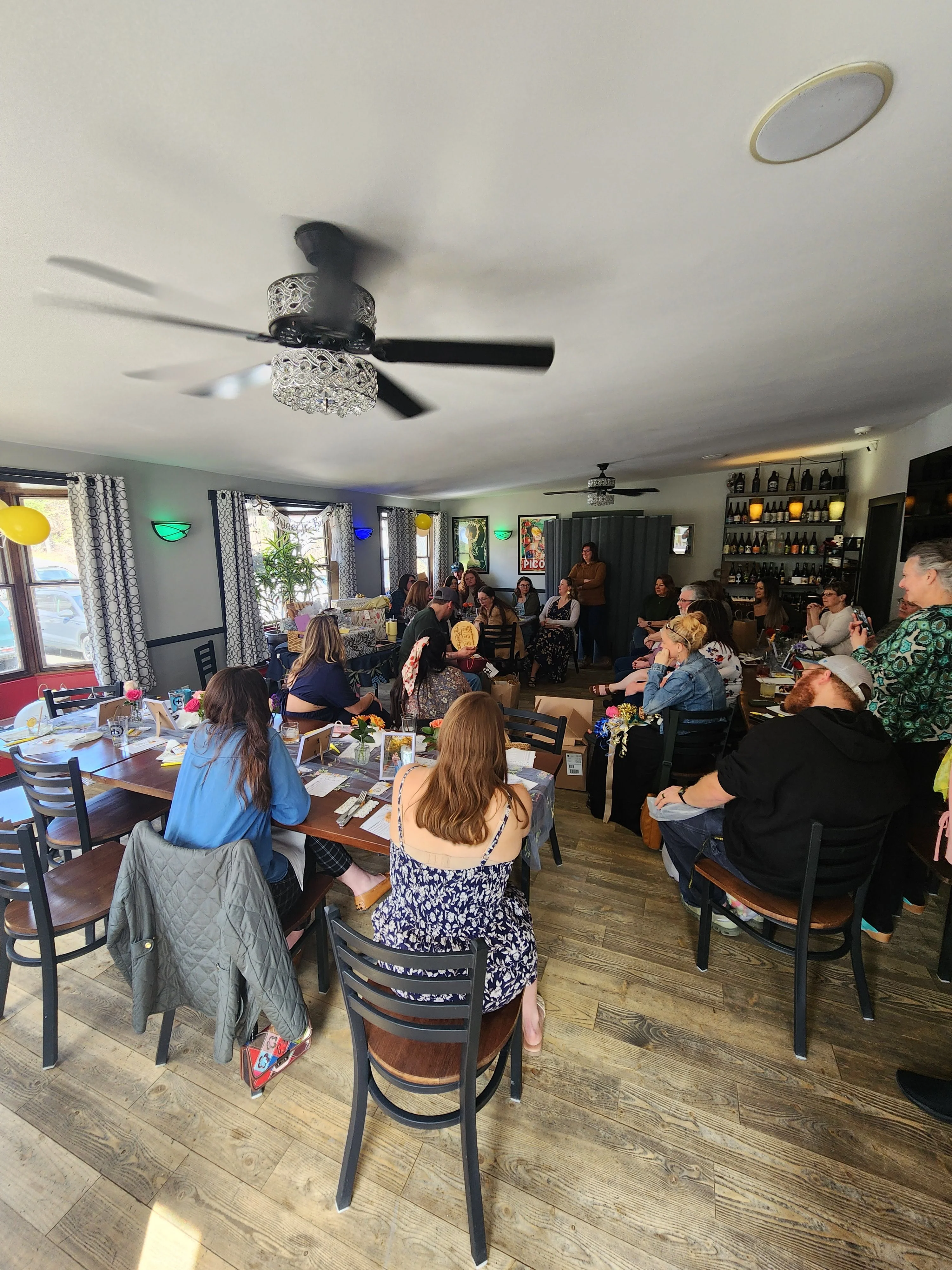 Busy restaurant dining room with guests, decorative ceiling fans, and bottle display