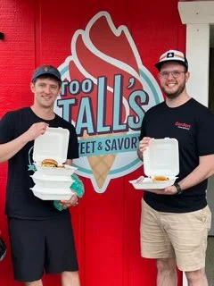 Two friends holding takeout burgers outside a vibrant ice cream shop