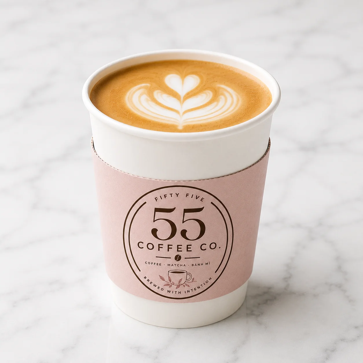 A take-out cup of coffee featuring a beautiful latte art design on top. The cup has a soft pink sleeve with a logo.