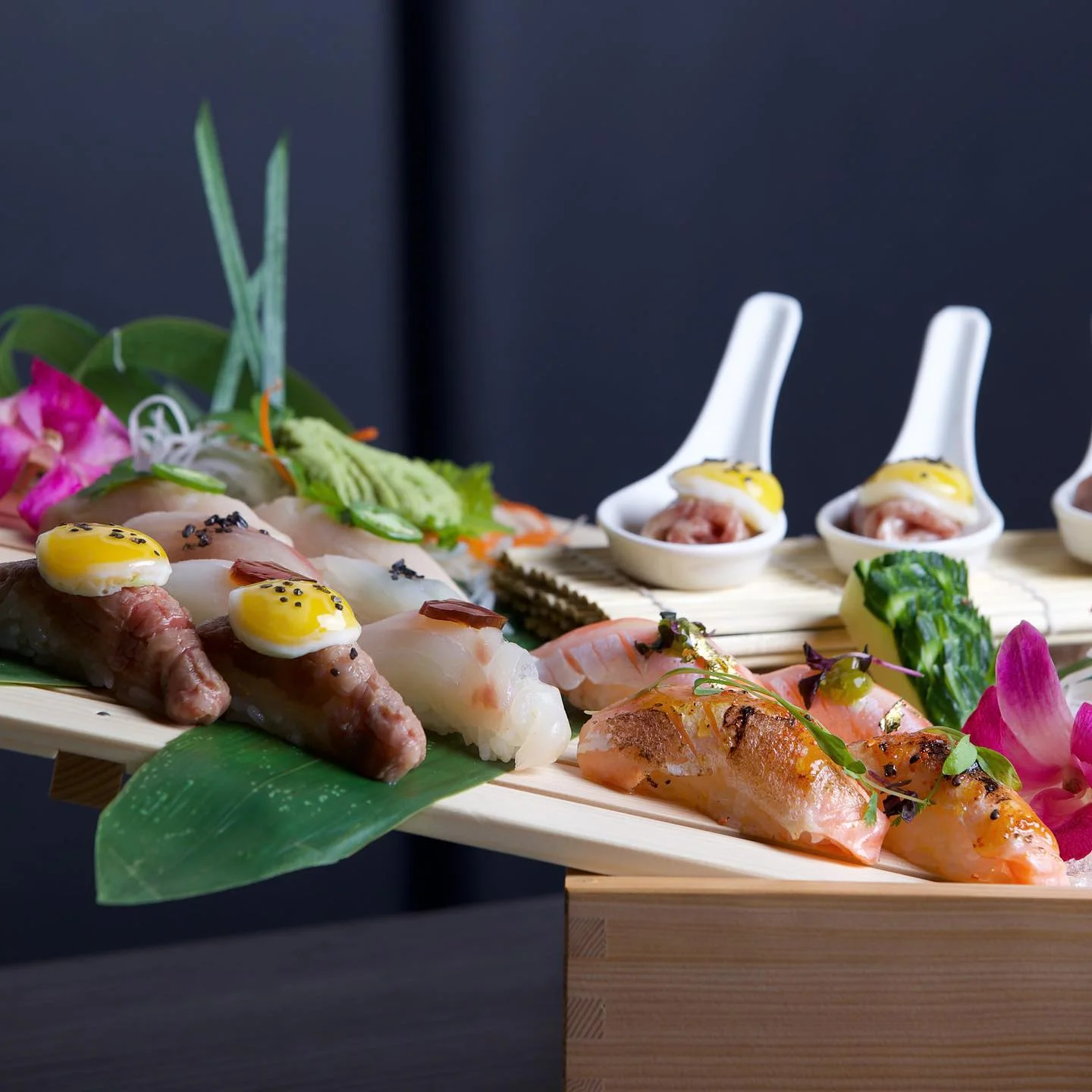 An elaborate sushi and sashimi platter presented on a wooden bridge tray, featuring seared meat nigiri topped with miniature fried quail eggs, seared salmon nigiri with gold leaf details, white fish nigiri, and individual tasting spoons with bite-sized appetizers in the background against a dark backdrop.