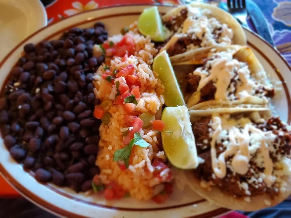A colorful plate featuring tacos topped with meat, cilantro, onions, and sauces, alongside sides of black beans and a vibrant rice mixture garnished with tomatoes and herbs.