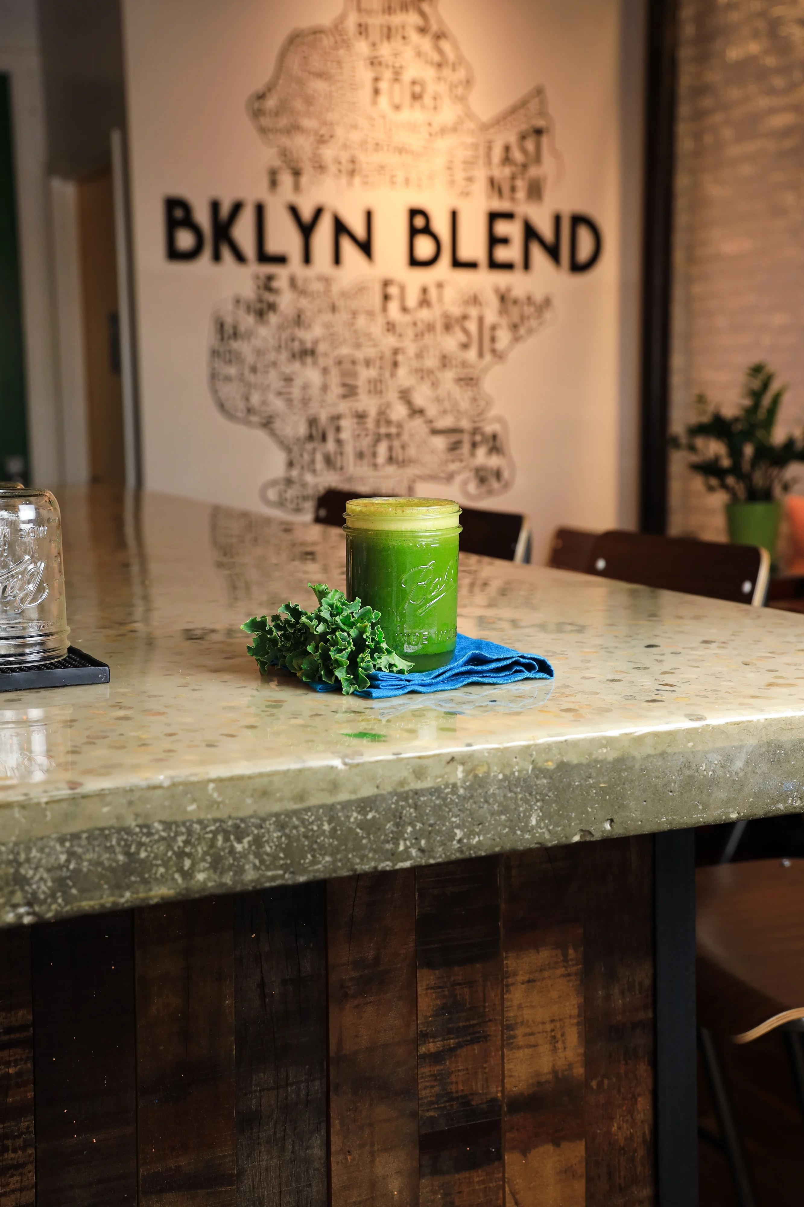 A green smoothie in a jar sits on a polished counter adorned with kale and a blue cloth. "BKLYN BLEND" is on the back wall, conveying a fresh, inviting cafe vibe.