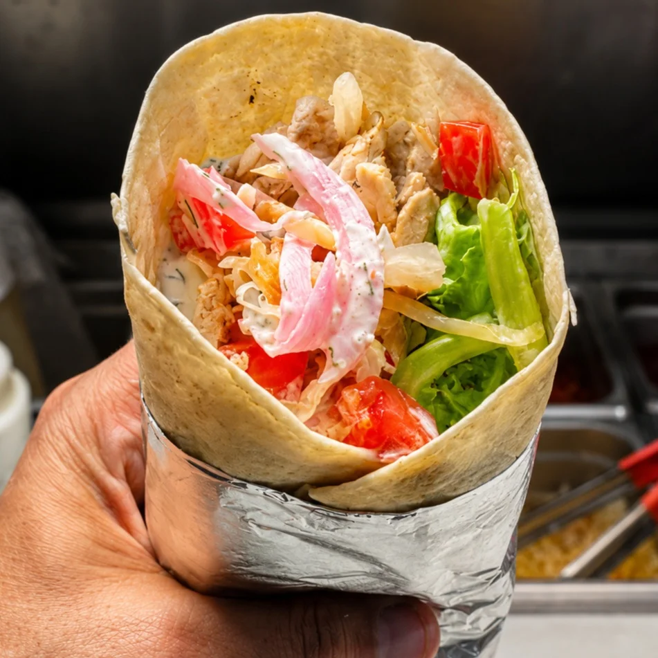 A hand holding a wrap filled with sliced meat, lettuce, tomatoes, and pink onions, wrapped in a tortilla.