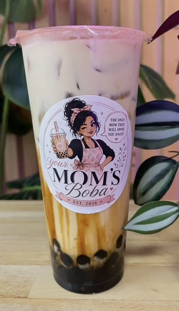 A transparent cup filled with a brown and creamy beverage, featuring tapioca pearls at the bottom. The cup has a colorful label with an illustration of a woman and text related to boba.