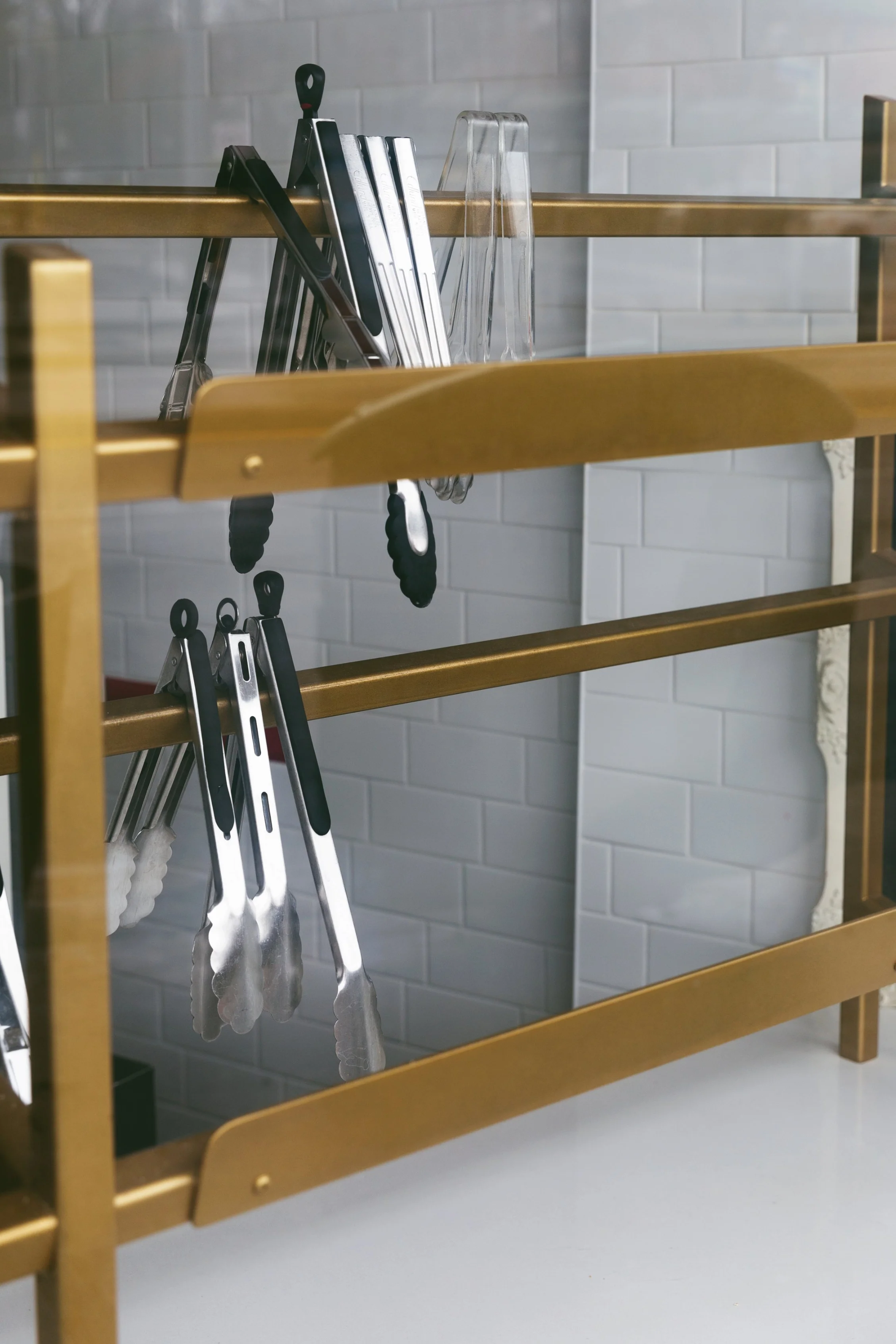 A gold and glass display rack featuring various kitchen tongs organized neatly in a row.