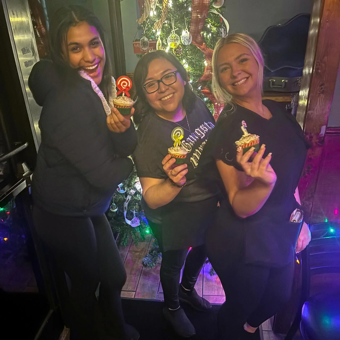 Three friends celebrating with cupcakes in front of a Christmas tree