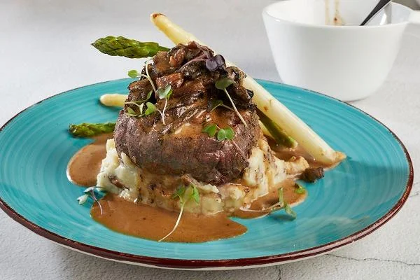 A gourmet dish featuring a tender beef filet topped with mushrooms and served on a bed of mashed potatoes, accompanied by asparagus and a rich gravy.