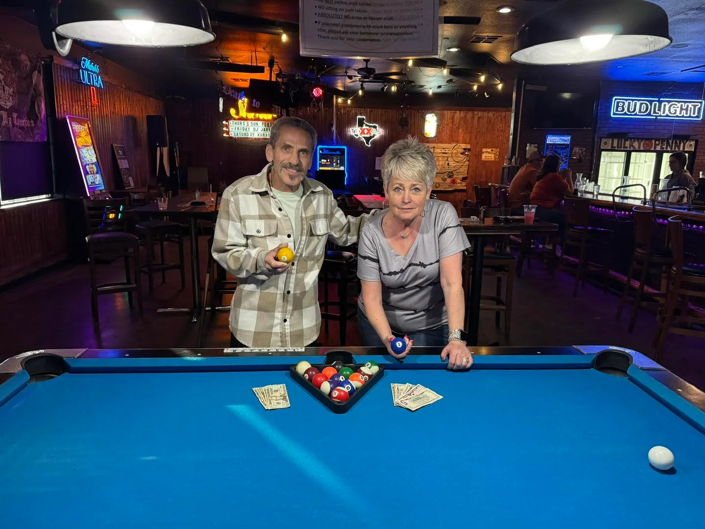 Two individuals posing by a pool table in a bar setting, one holding a yellow ball and the other holding a blue ball. Money is seen on the table.