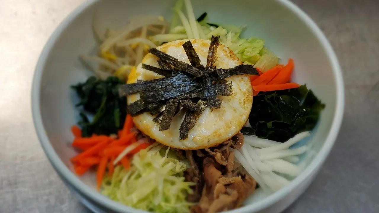Colorful Korean bibimbap bowl with rice, assorted vegetables including spinach, carrots, bean sprouts, and cabbage, topped with a fried egg and crispy seaweed strips, served at Sushi Kim 2 restaurant