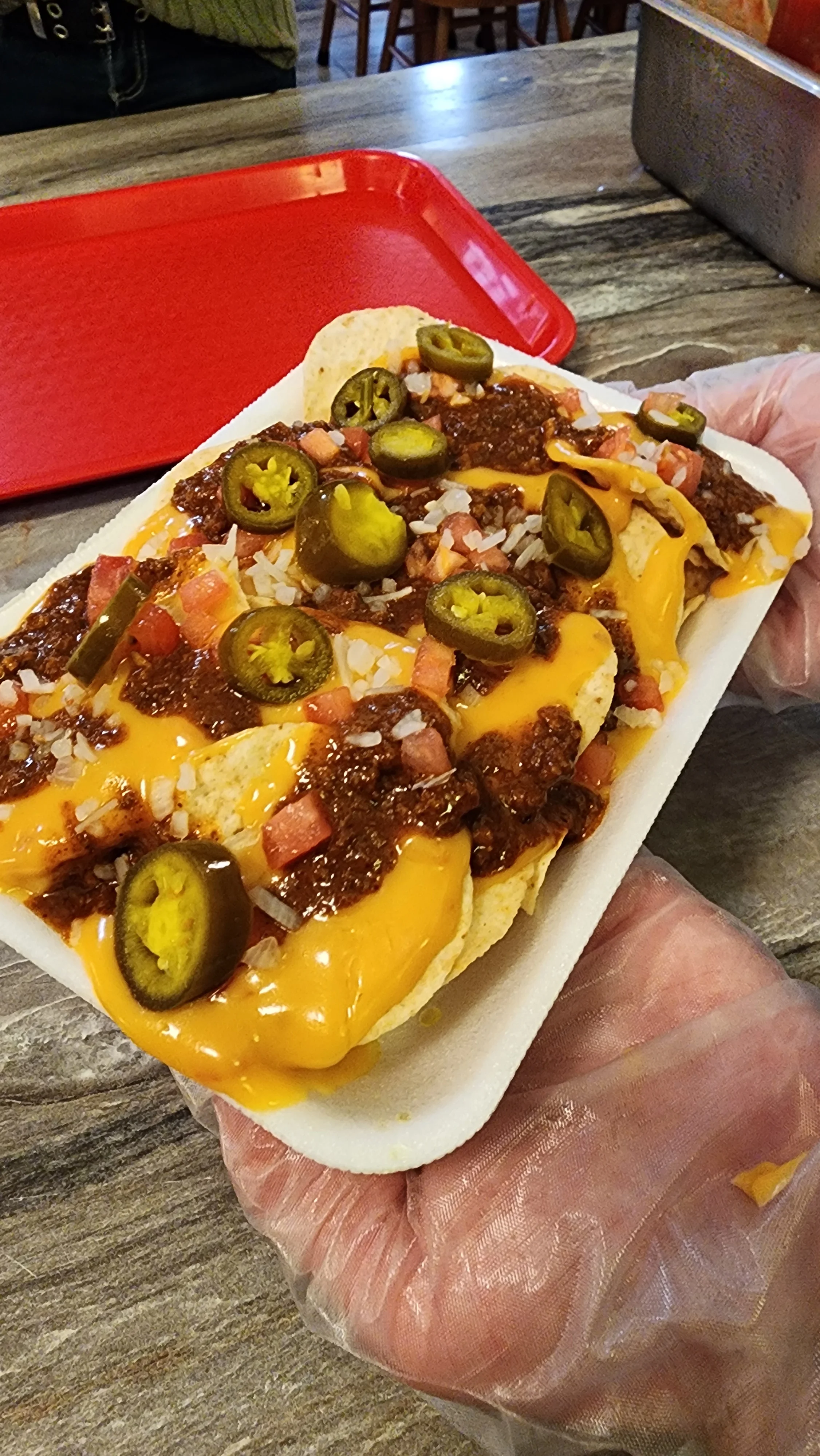 A plate of nachos topped with cheddar cheese, chili, diced tomatoes, onions, and jalapeños, held in a gloved hand.