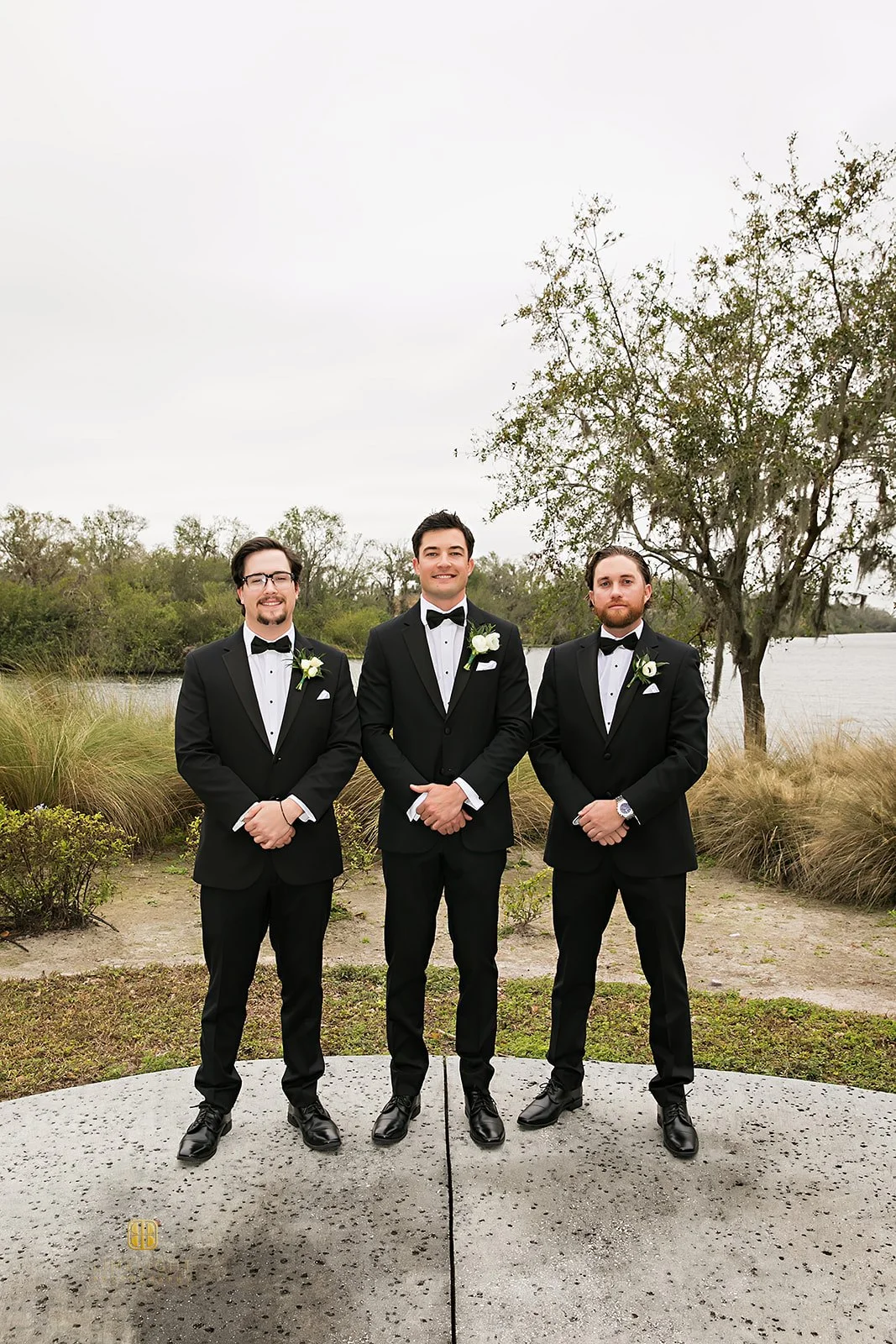 Three men in tuxedos posing outdoors by a lake