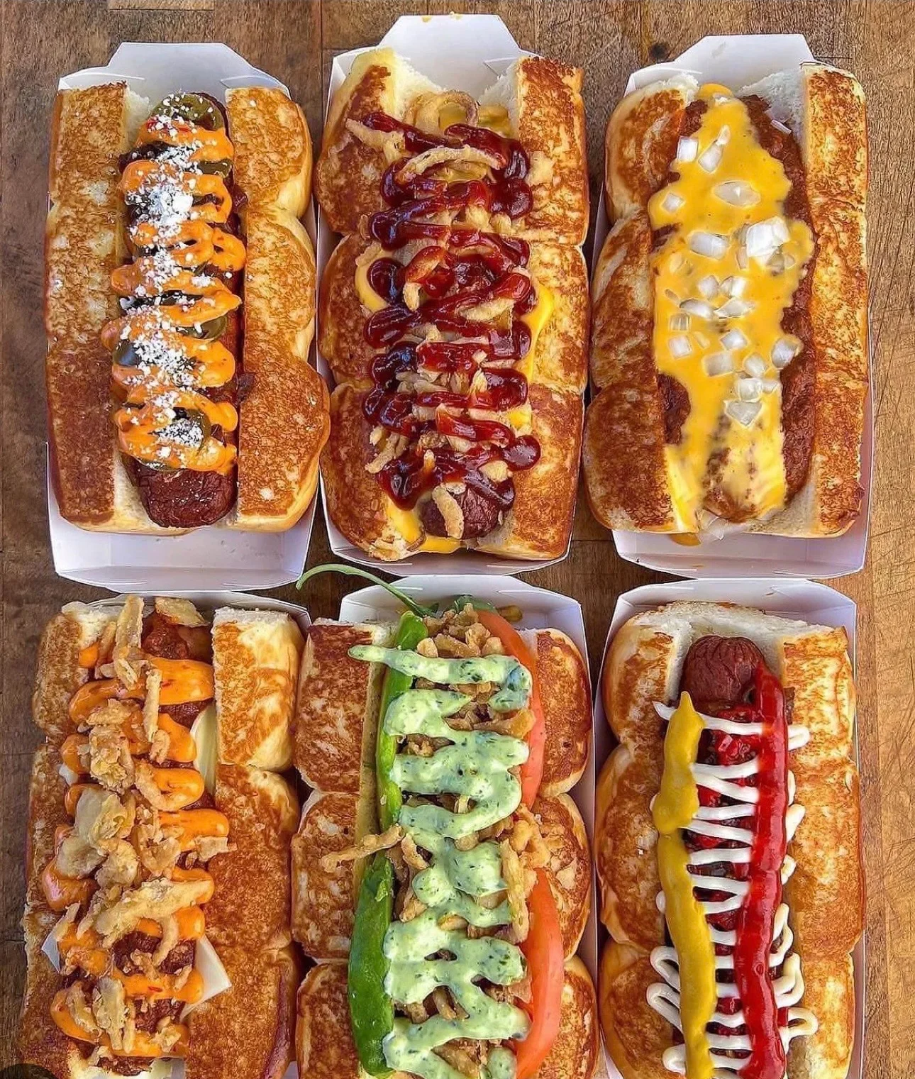 Six gourmet hot dogs with various toppings in white trays.