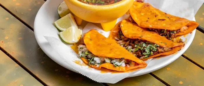 A plate of tacos with golden, crispy tortillas filled with meat, onions, and garnished with cilantro. Accompanied by lime wedges and a bowl of salsa.