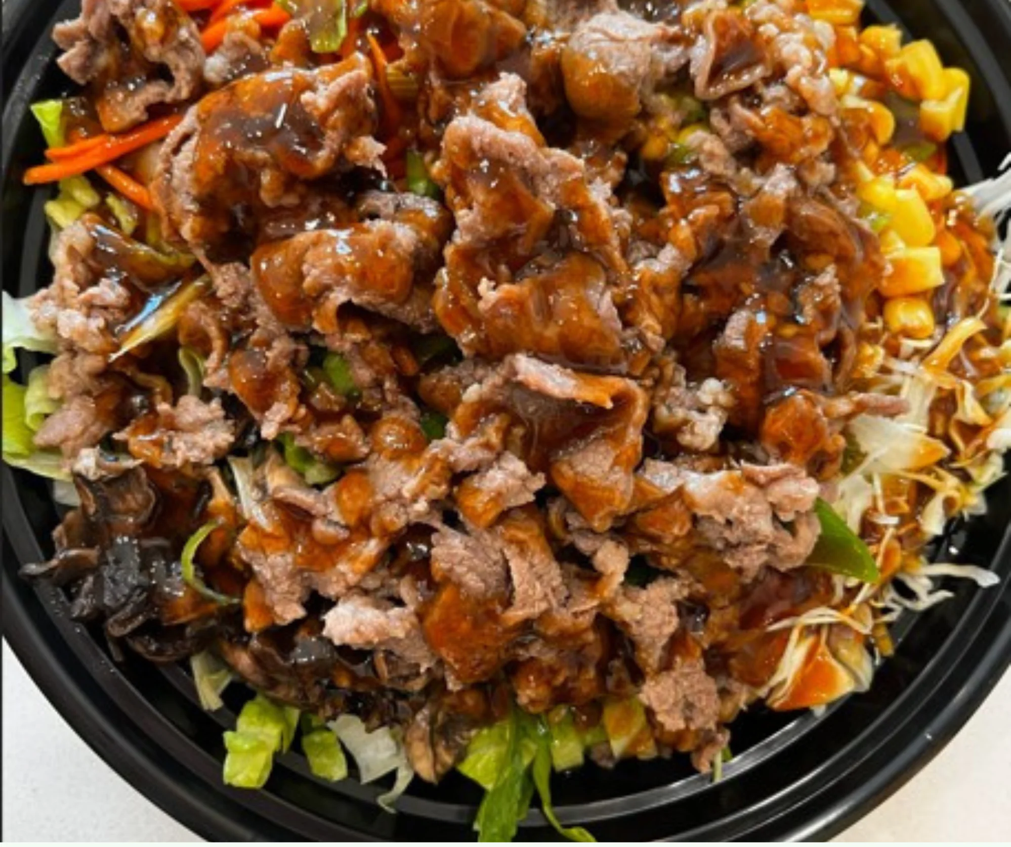 A colorful bowl filled with shredded lettuce, corn, carrots, and strips of beef drizzled with a savory sauce.