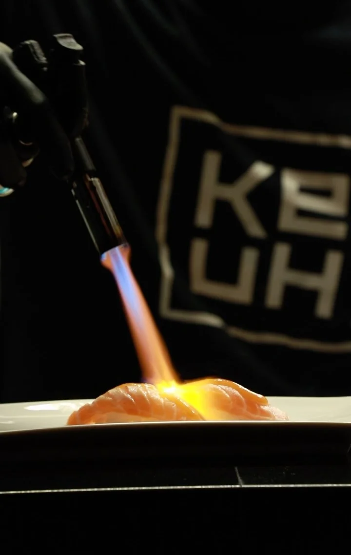 A chef using a kitchen torch to sear fresh salmon nigiri on a white plate.