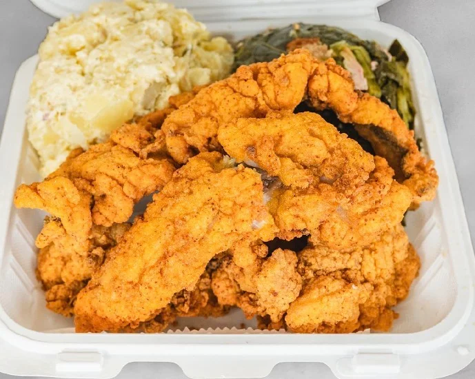 A serving of fried fish fillets accompanied by sides of mashed potatoes and collard greens in a takeout container.