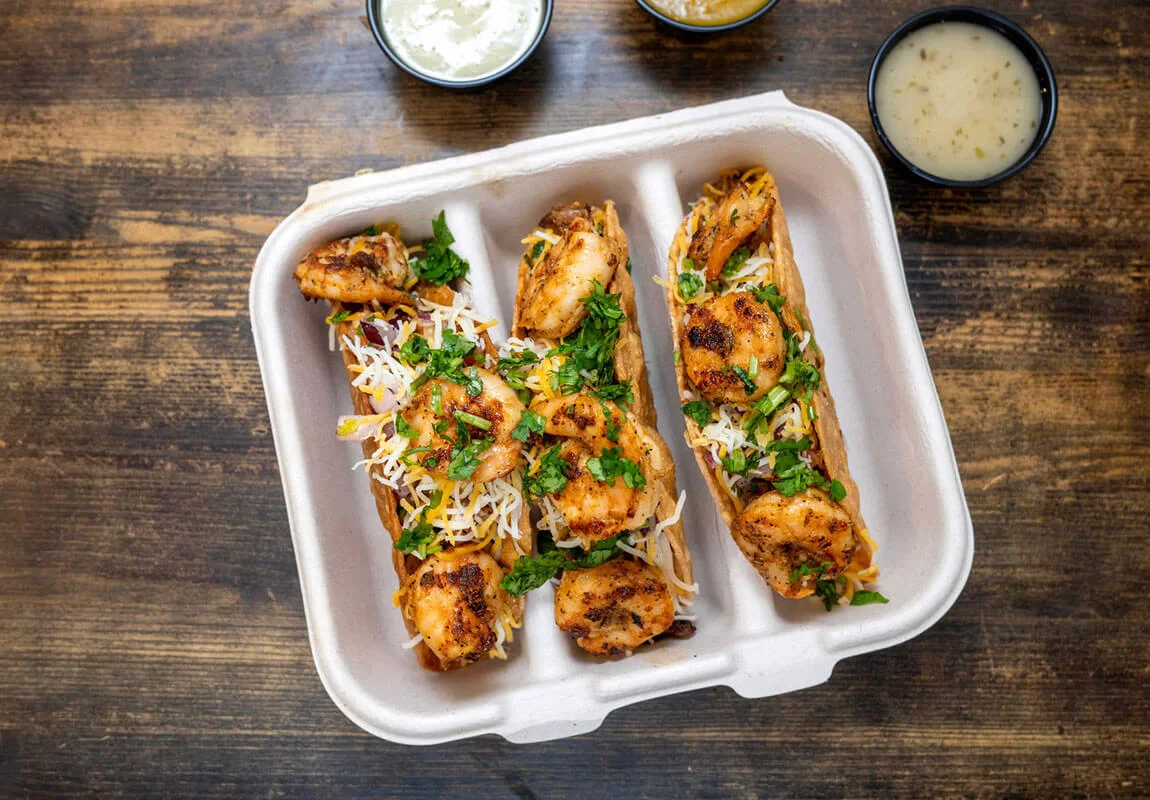 Three shrimp tacos served with fresh herbs and cheese in a takeout container, accompanied by dipping sauces.