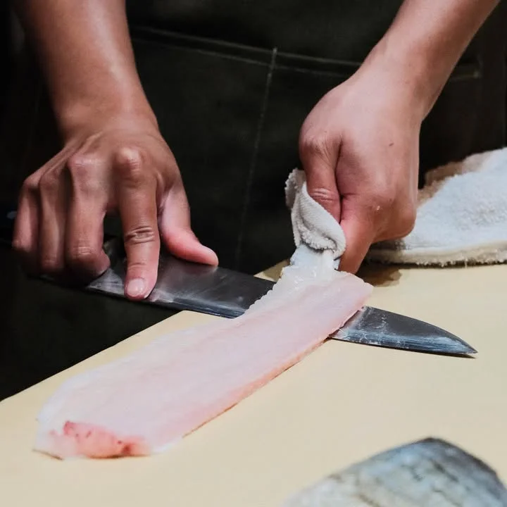 A close-up view of hands skillfully slicing a piece of fish with a sharp knife on a wooden cutting board.