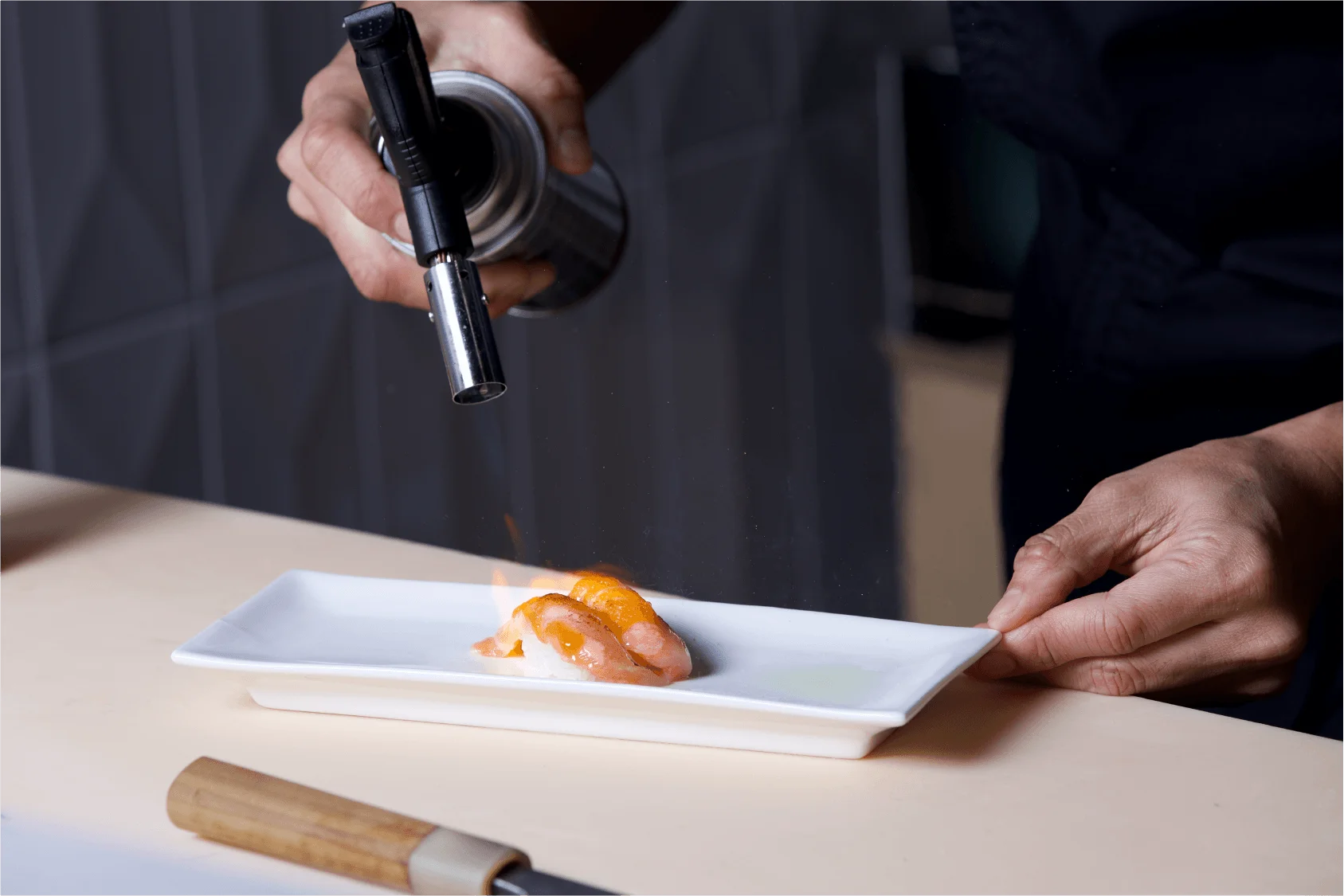 A chef using a kitchen blowtorch to sear pieces of salmon nigiri sushi on a rectangular white plate.