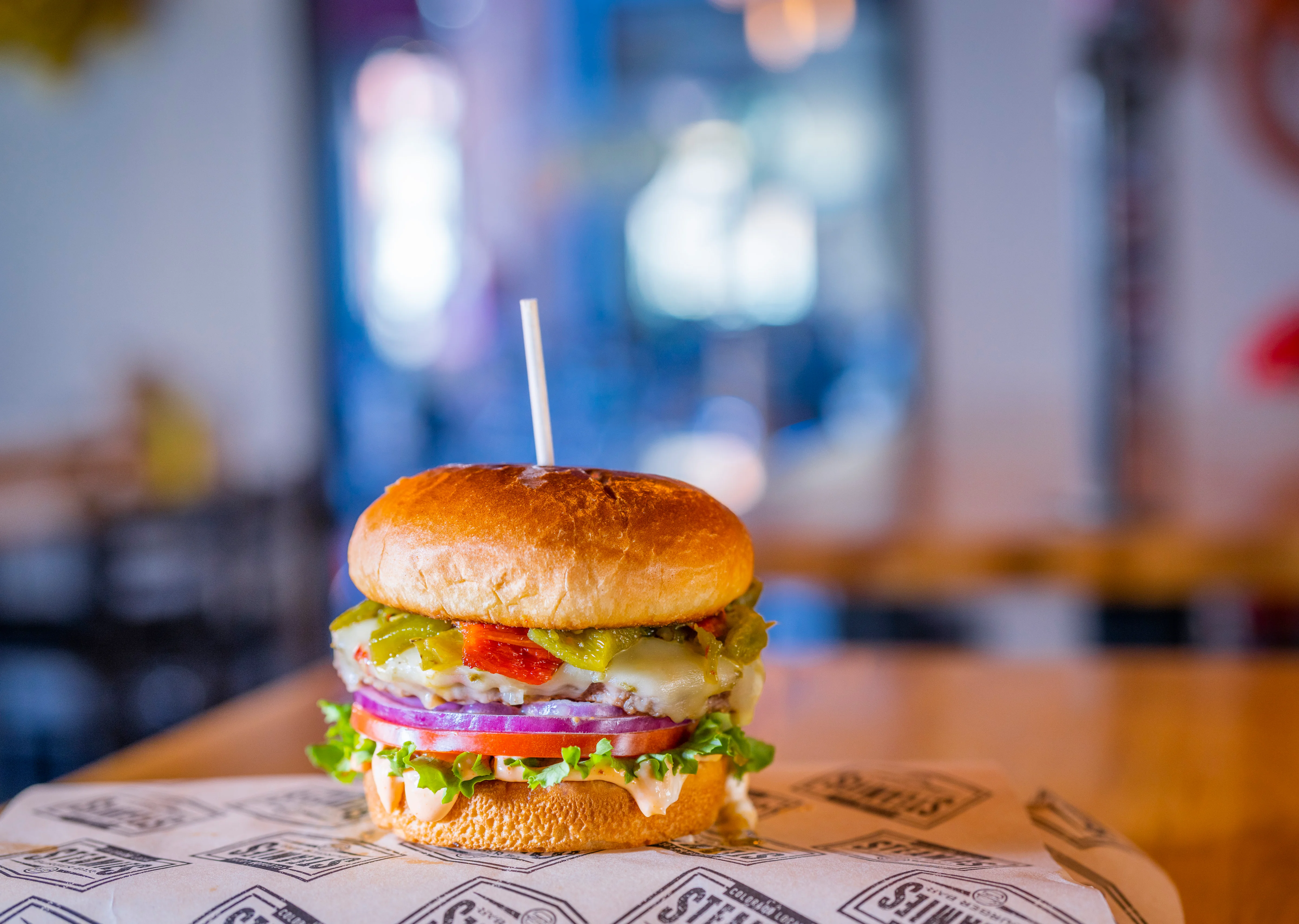 A loaded hamburger featuring layers of lettuce, tomato, onion, pickles, and melted cheese, served on a toasted bun and sitting on patterned paper.