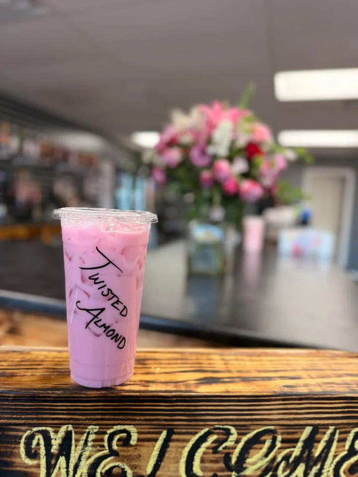 A pink drink with ice, prominently displayed on a wooden surface with flowers in the background.
