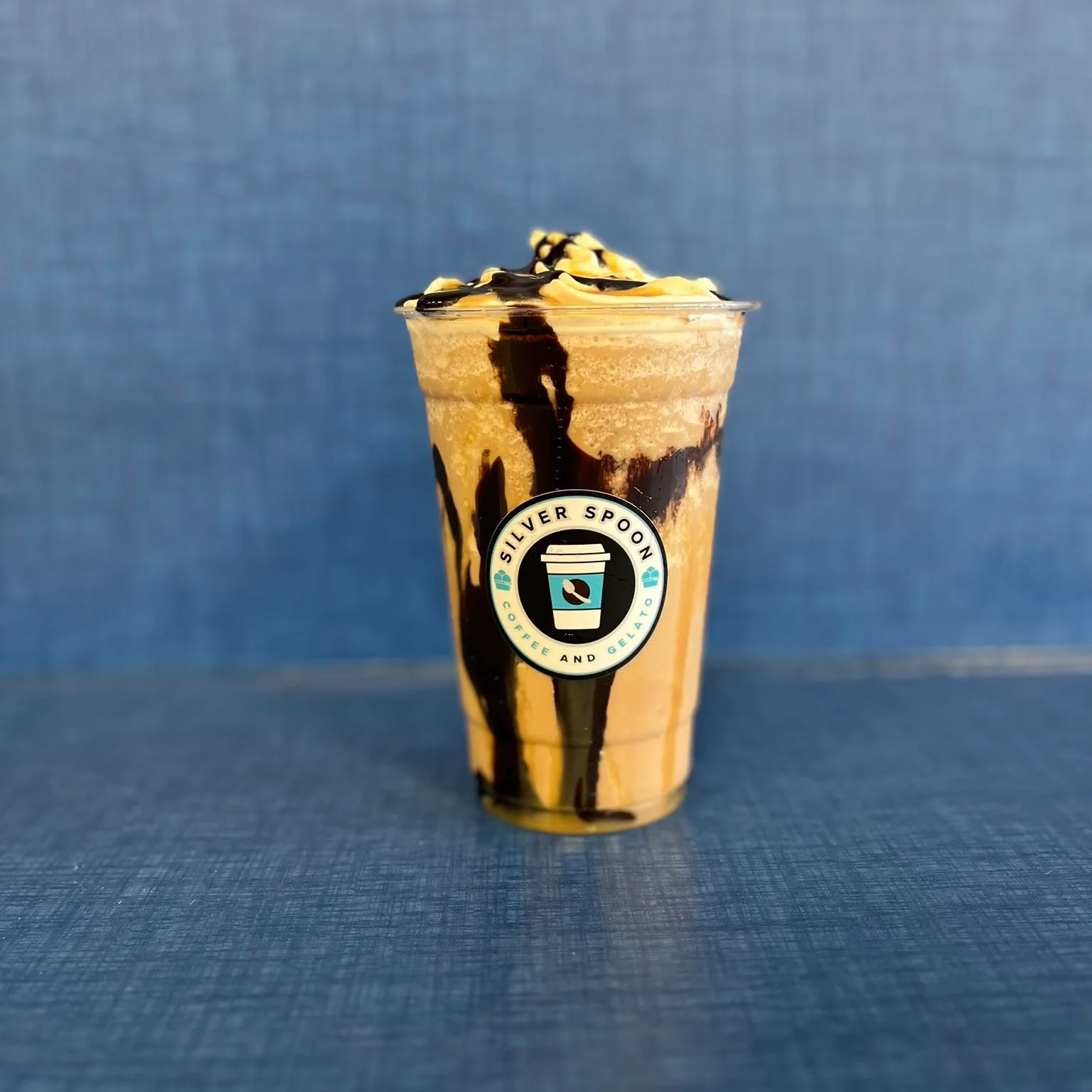 A plastic cup of a coffee beverage with whipped cream, featuring the 'Silver Spoon' logo, placed on a wooden table.