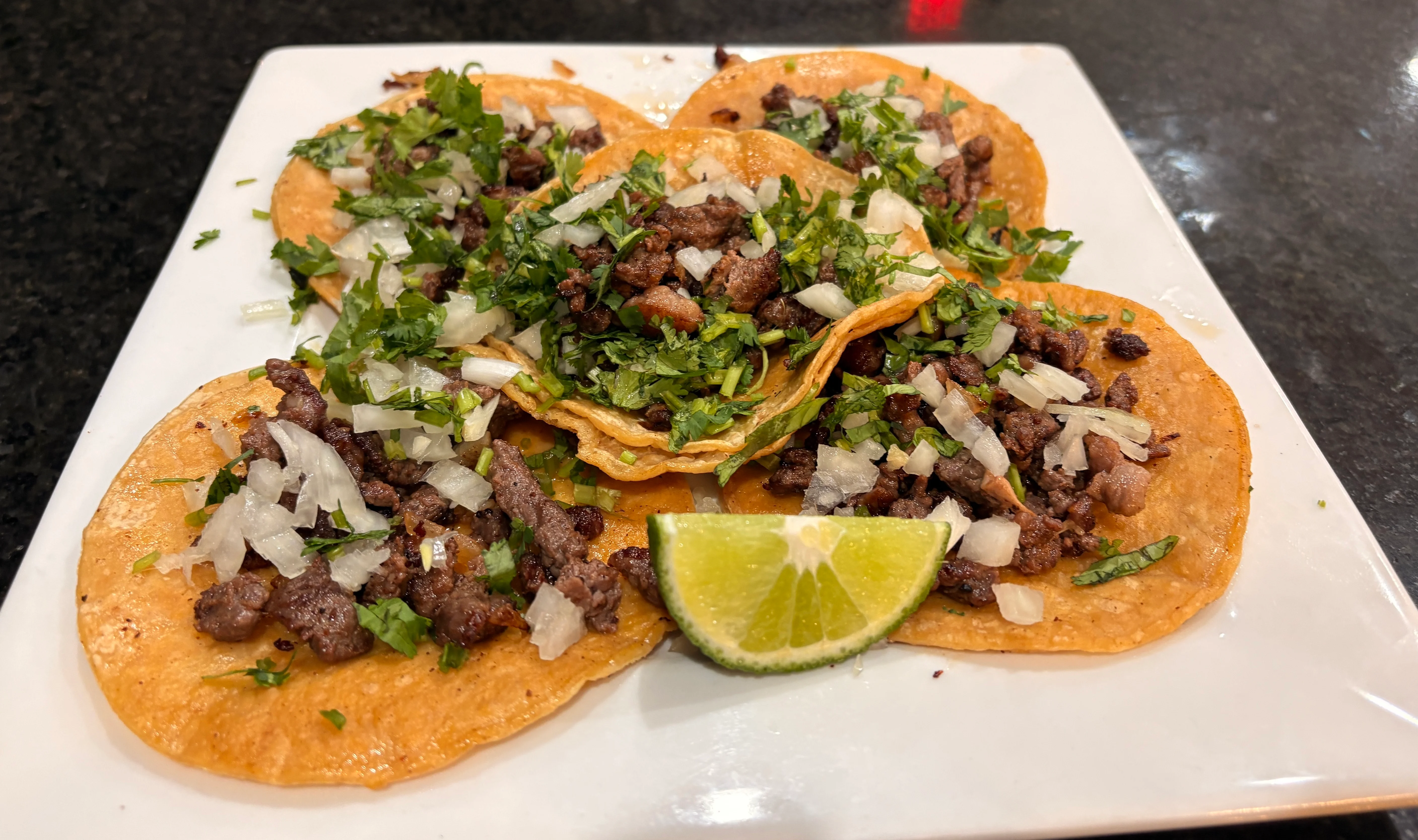 A plate of soft corn tortillas topped with seasoned meat, fresh cilantro, and diced onions, accompanied by a lime wedge.