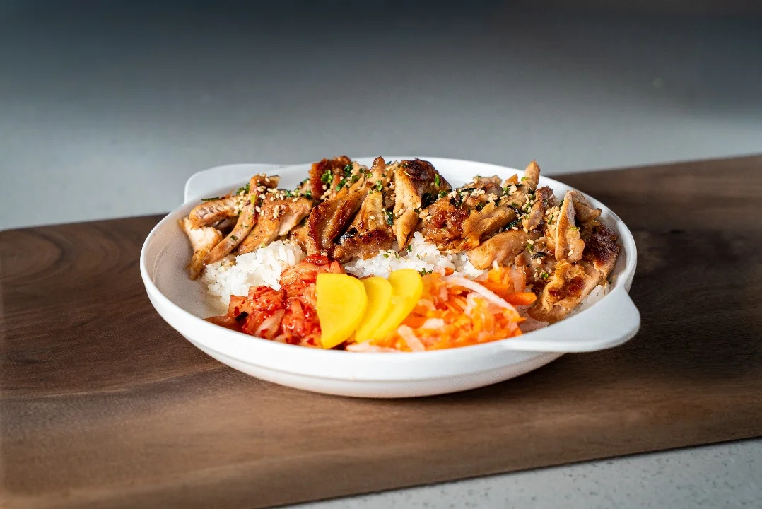 A rice bowl topped with grilled chicken, garnished with sesame seeds, and accompanied by pickled vegetables and kimchi.