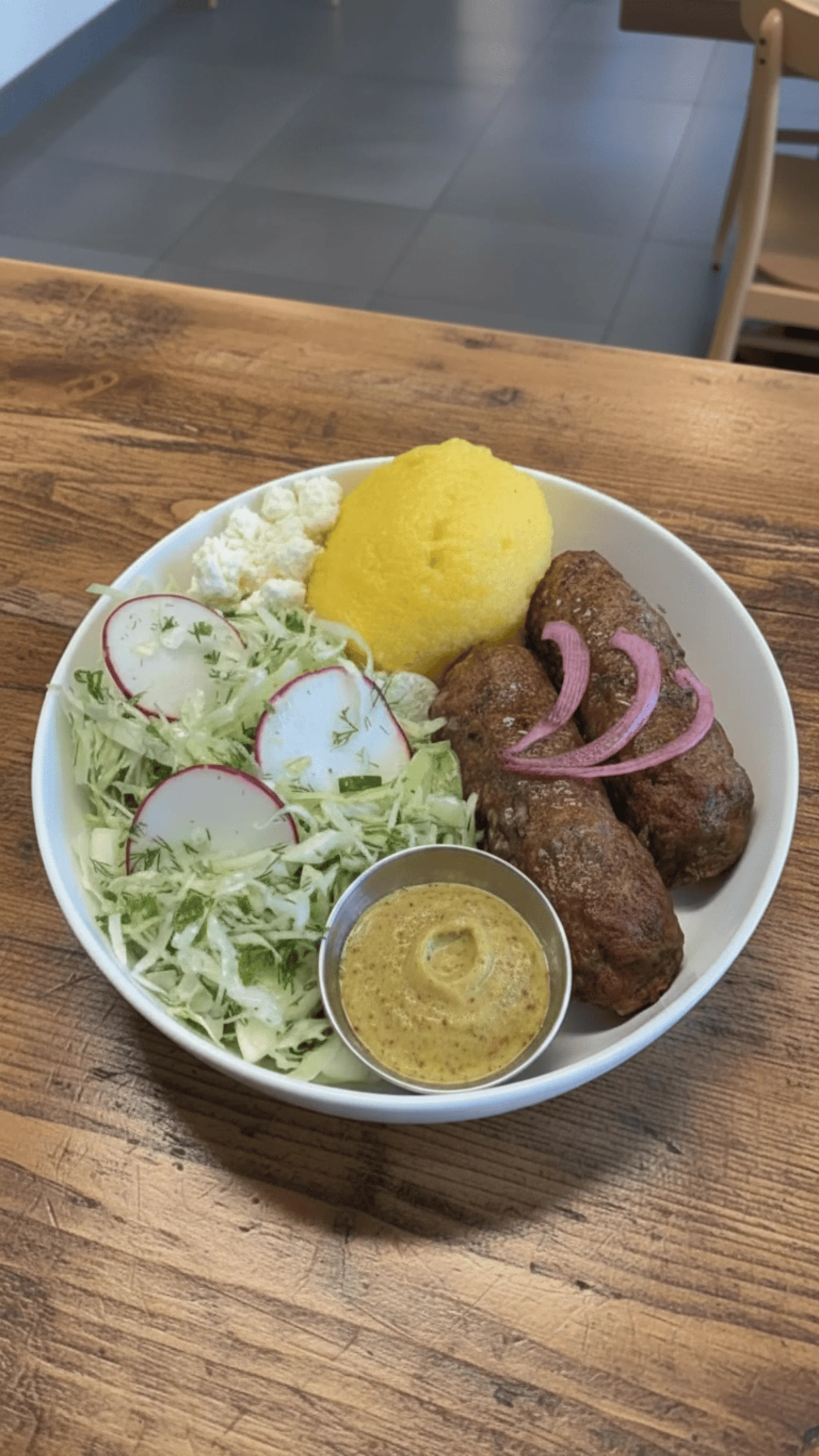 A plate featuring grilled skewers, polenta, a salad with radishes, and a small bowl of mustard or sauce.