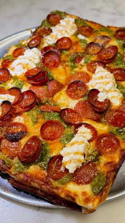 A heart-shaped pizza topped with cherry tomatoes, olives, and green peppers, being sliced by a hand with a fork and knife. There is a candle in the background.