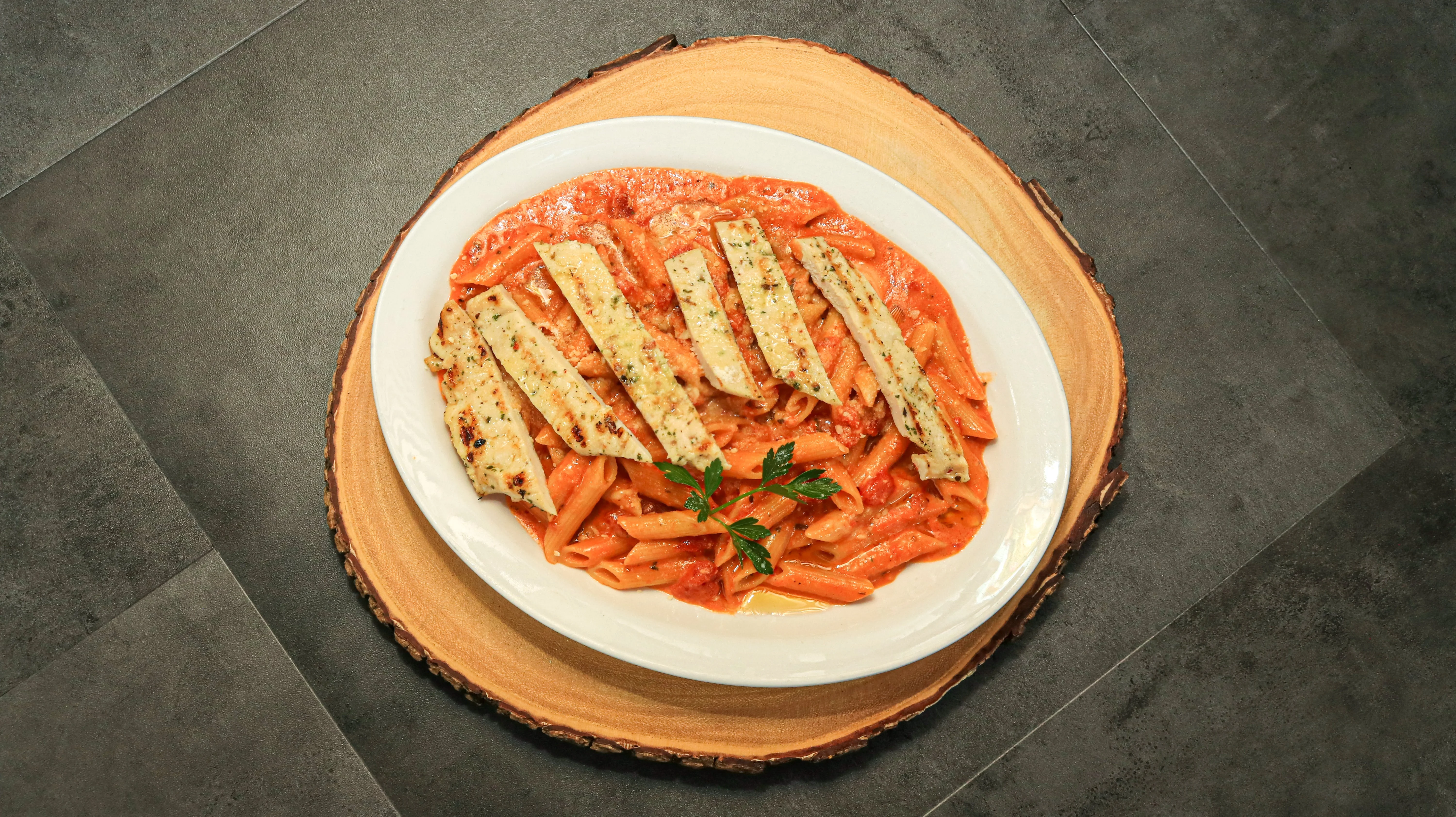 A plate of pasta with a creamy tomato sauce, topped with grilled strips of chicken and a sprinkle of herbs, displayed on a wooden platter.