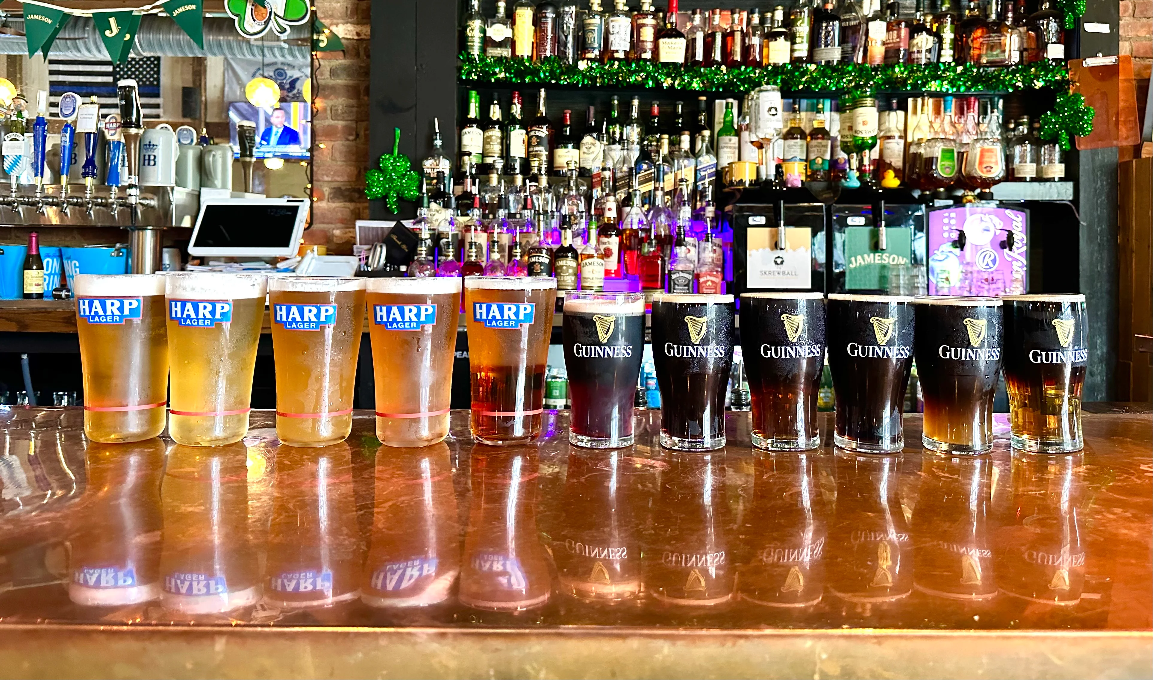 A variety of beers arranged on a bar counter, including Harp Lager and Guinness. The background features a shelf filled with various liquor bottles.