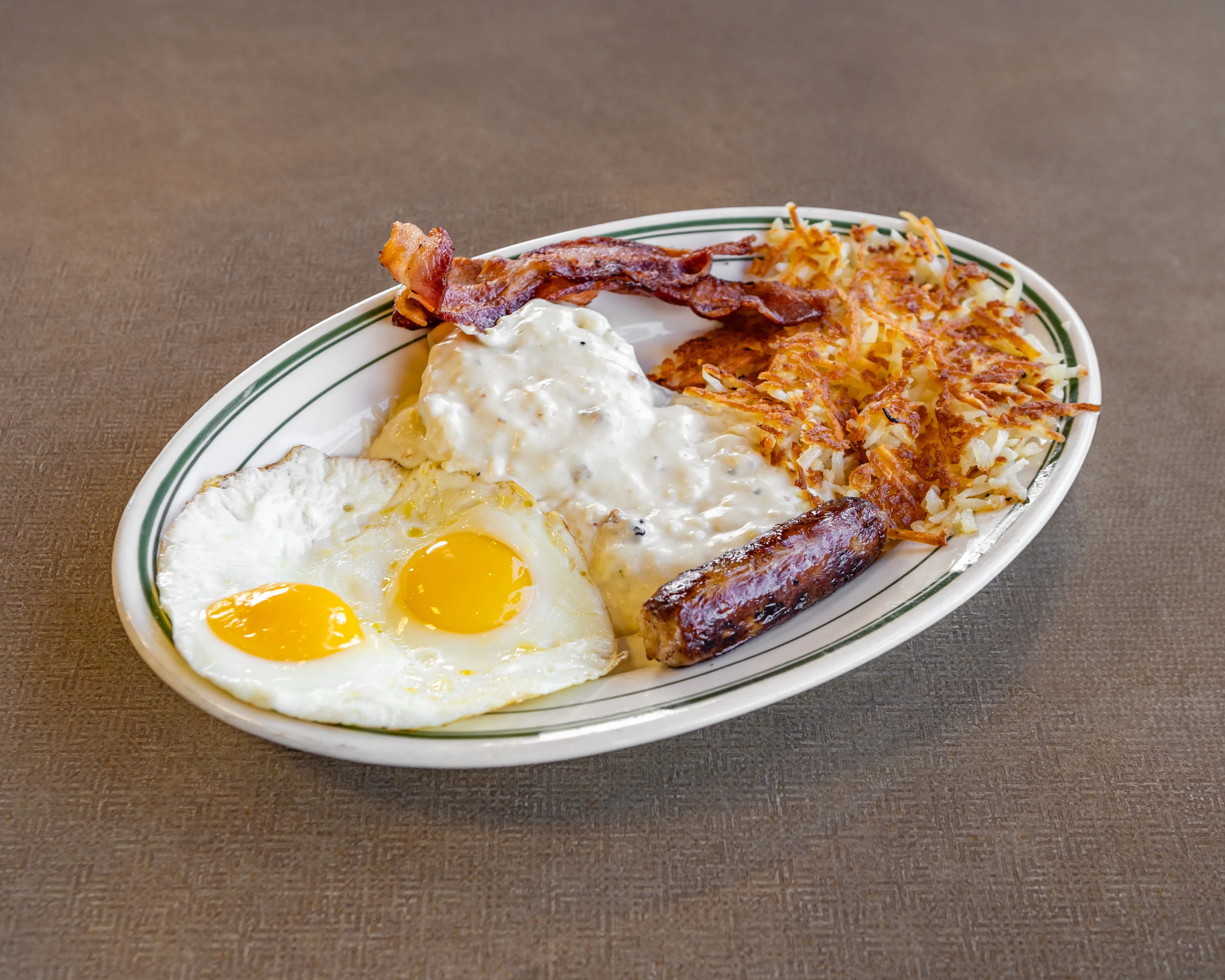 A breakfast plate featuring two fried eggs, sausage, bacon, and hash browns topped with gravy, served on a white plate.