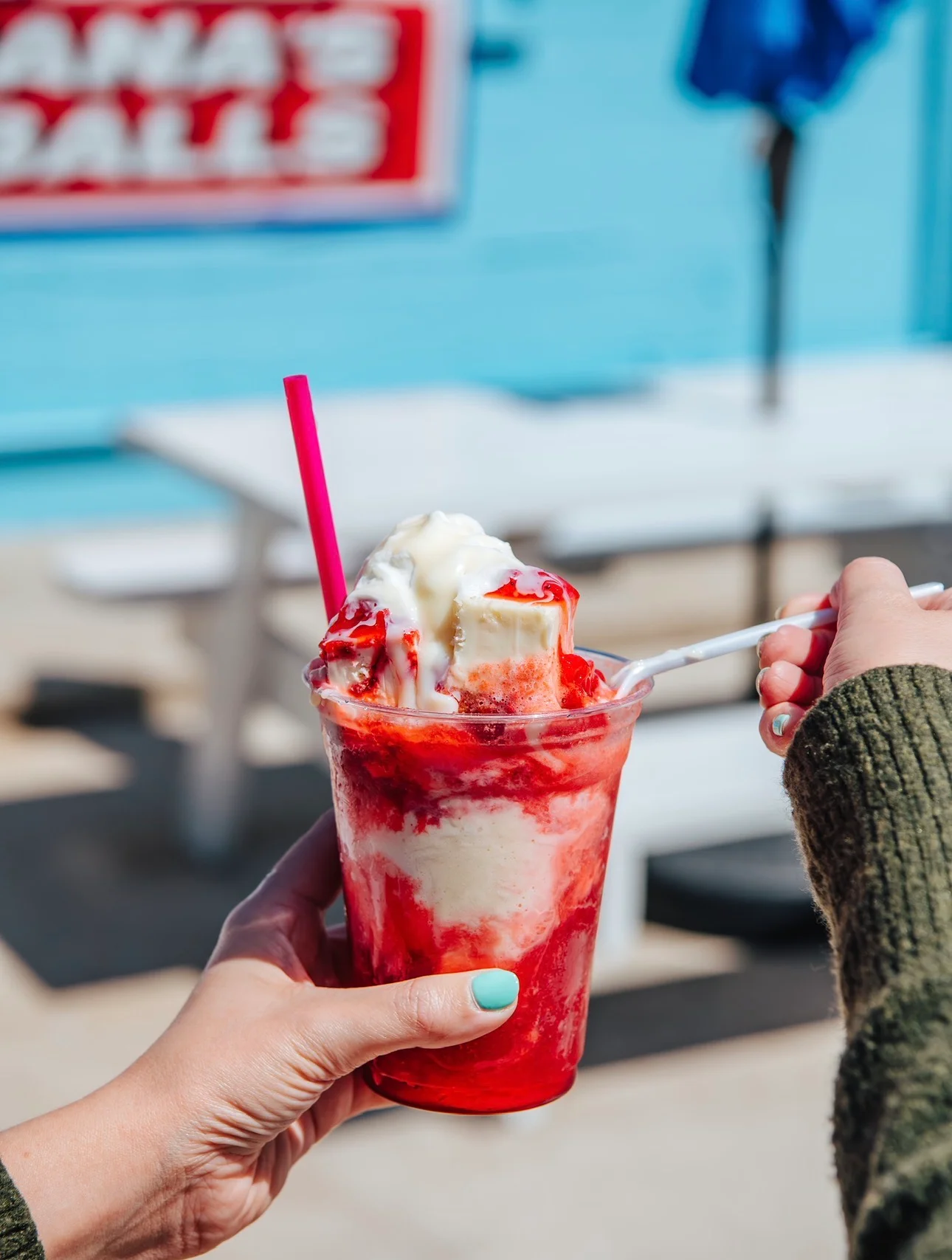 A cup of red snow cone topped with creamy syrup, being scooped with a spoon.